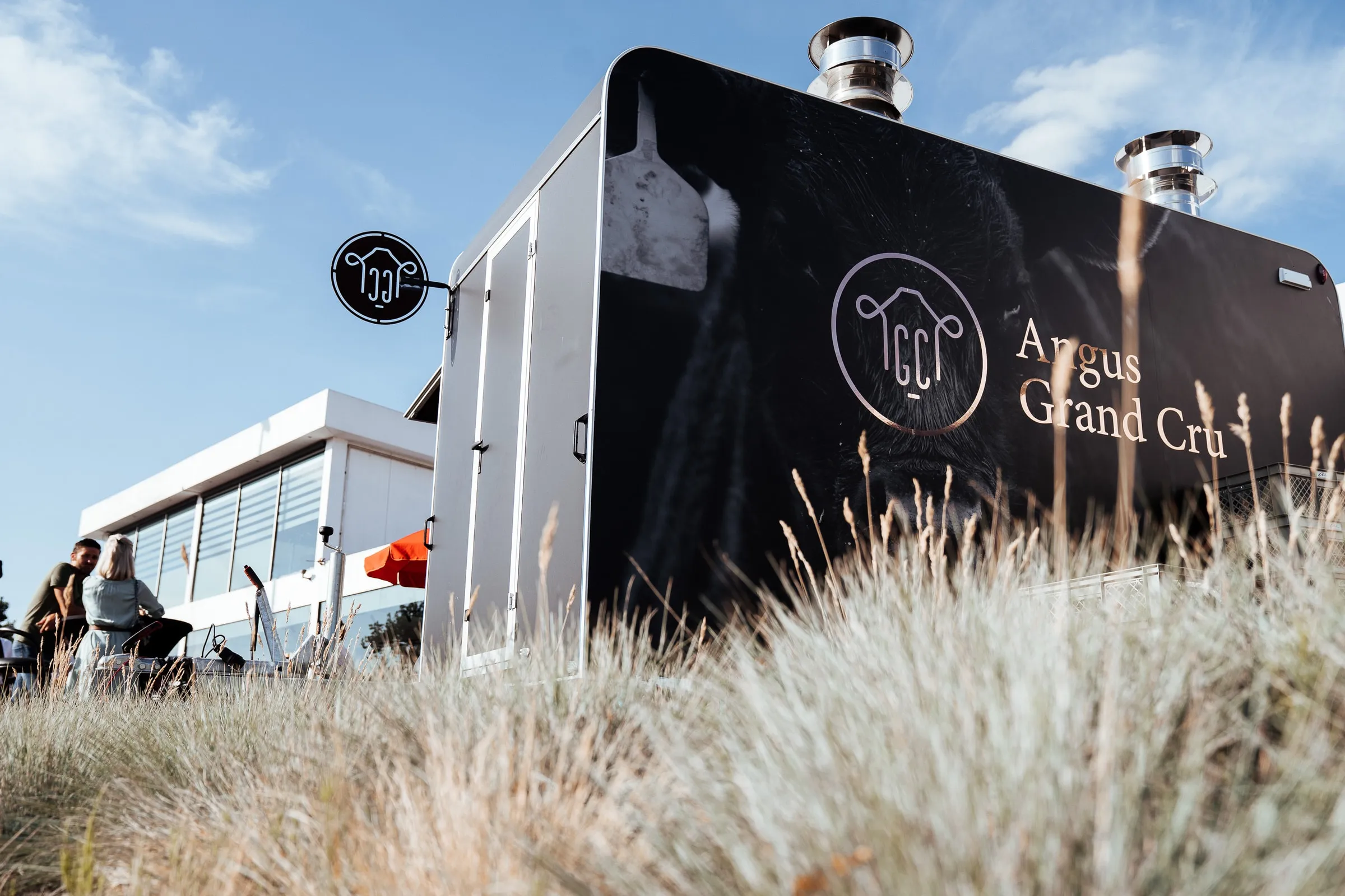 De zwarte foodtruck van Angus Grand Cru met het logo, in het hoge gras op een zonnig event